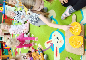 Na desce na dywanie rozkrojona dynia. Dzieci kolejno oglądają i dotykają ziarenka.